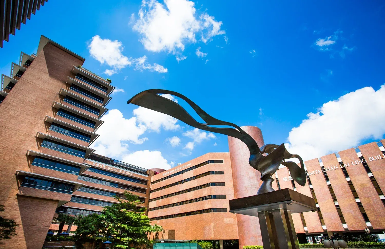 Modern multi-story building with brown brick exterior and glass balconies, with a large abstract metal sculpture in the foreground, under a blue sky with white clouds.